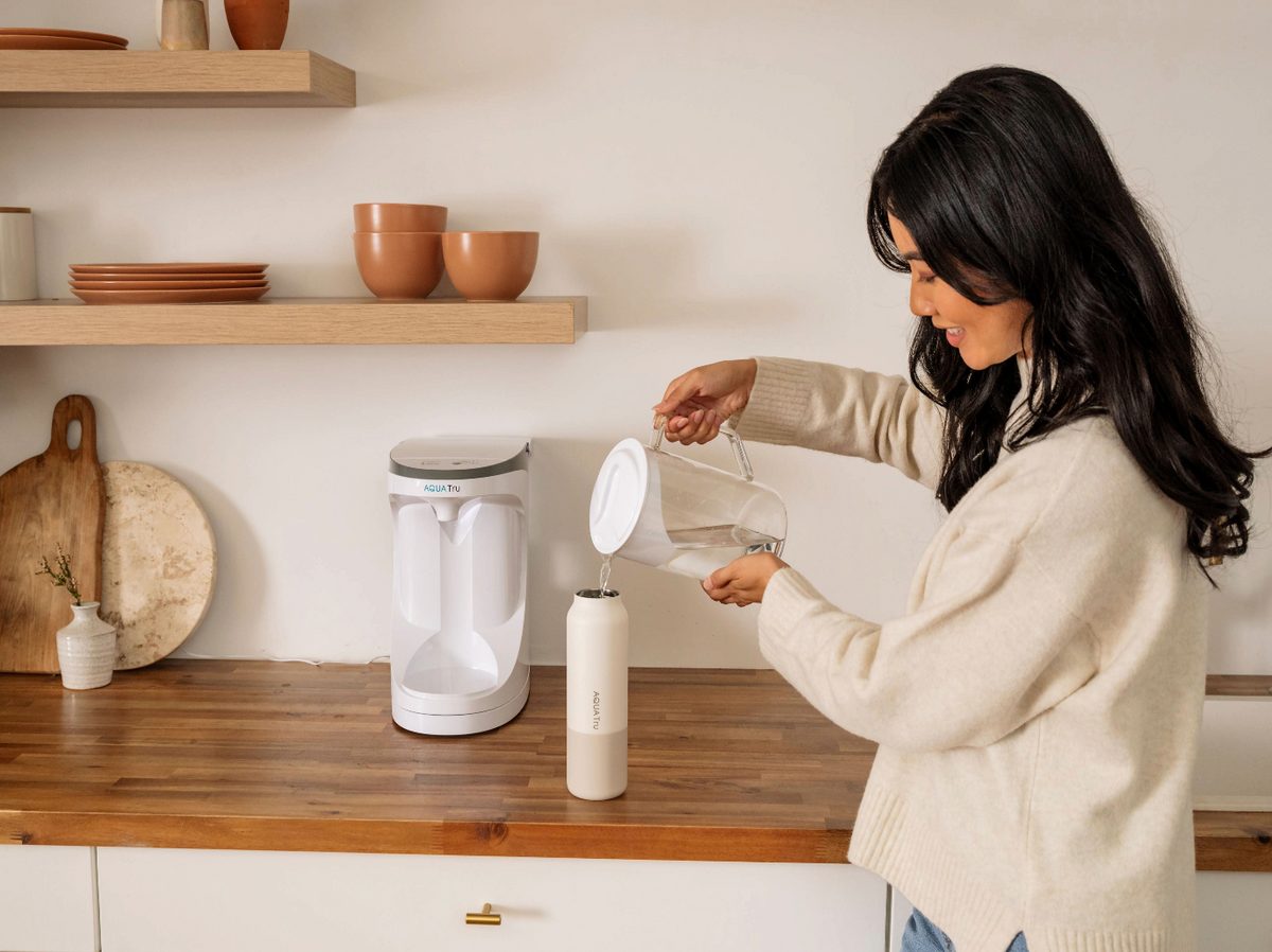 A woman pours water from a carafe into a sleek white water bottle on a wooden countertop with kitchenware.