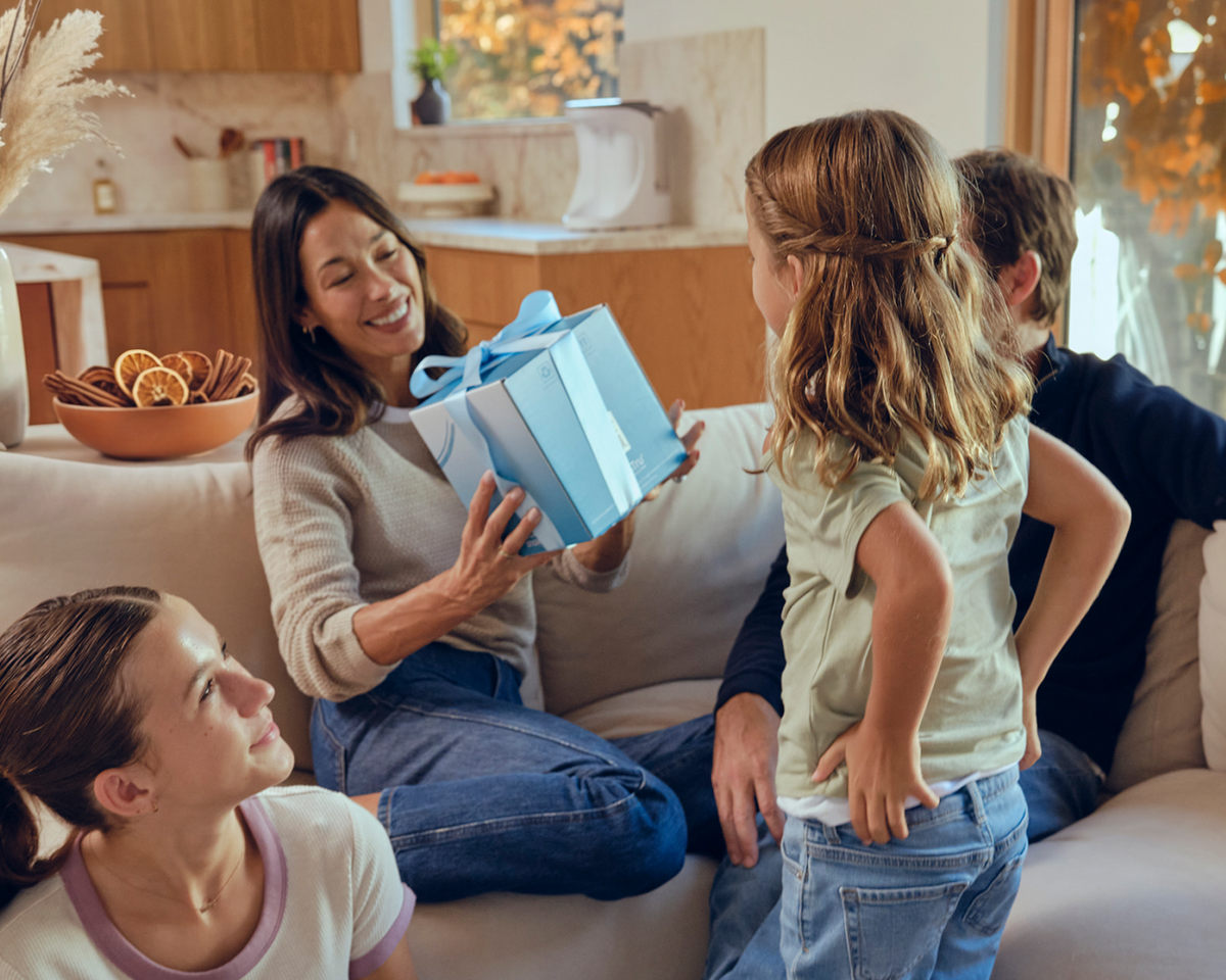 A woman hands a blue gift box to a girl while two others watch, all sitting on a cozy couch in a warm living room.
