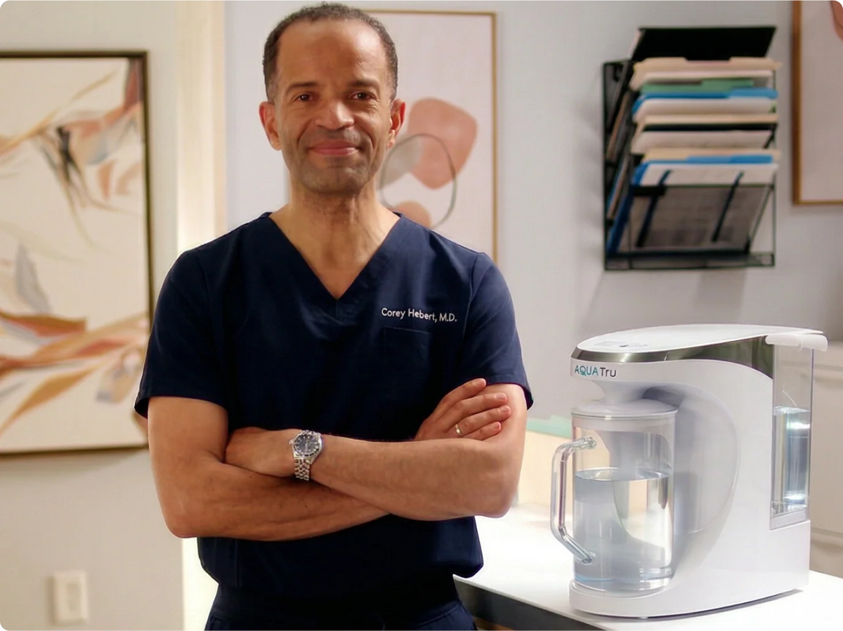 Dr. Corey Herbert stands confidently with arms crossed beside an AQUA Tru water filtration system.