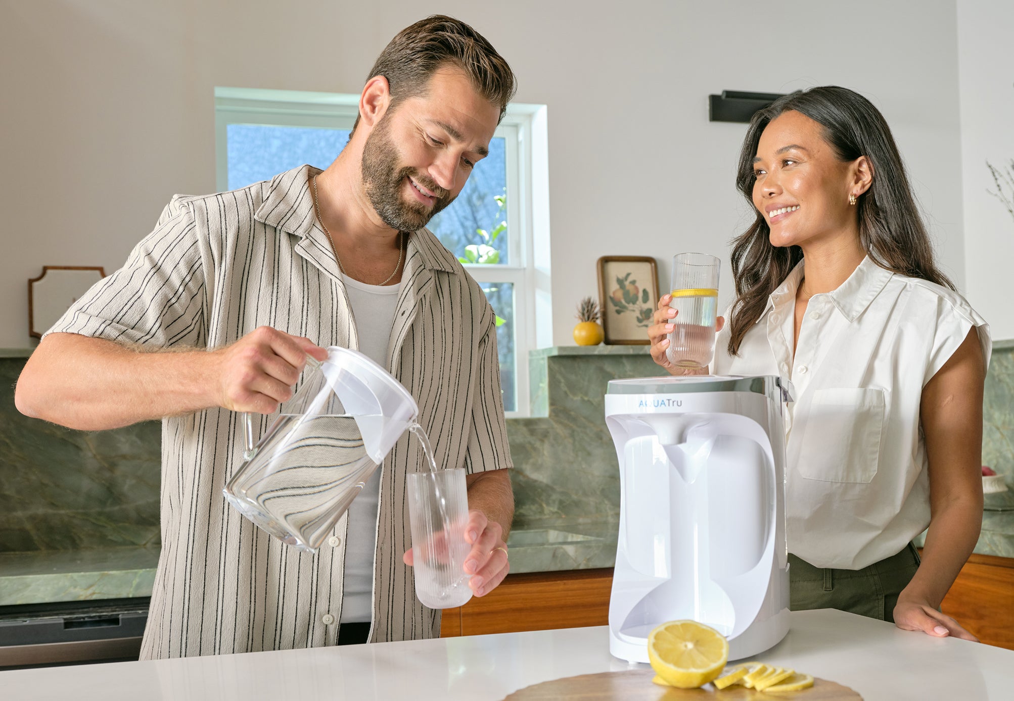 A man pours water from a carafe into a glass while a woman smiles, standing beside a countertop with a lemon.