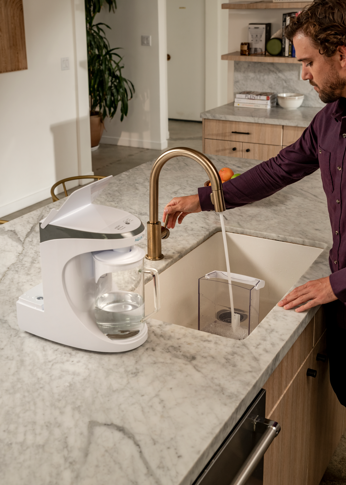 A man refills a carafe tank at a kitchen sink, with a coffee maker on the counter and a marble surface.