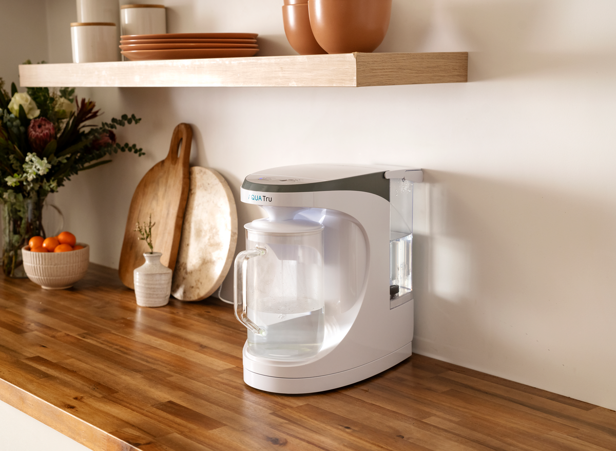 AQT Carafe on a wooden countertop with kitchenware, plants, and fruit in the background.