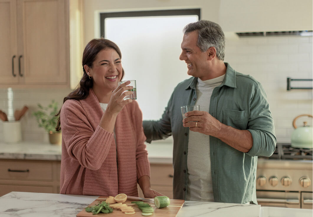 A couple smiles and toasts with glasses in a bright kitchen, surrounded by fresh fruits and vegetables on a cutting board.
