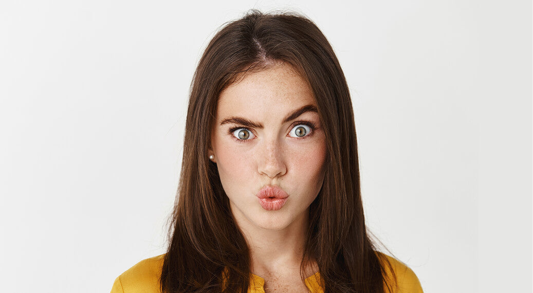 Young woman with long brown hair making a playful pouty face, wearing a yellow top against a white background.