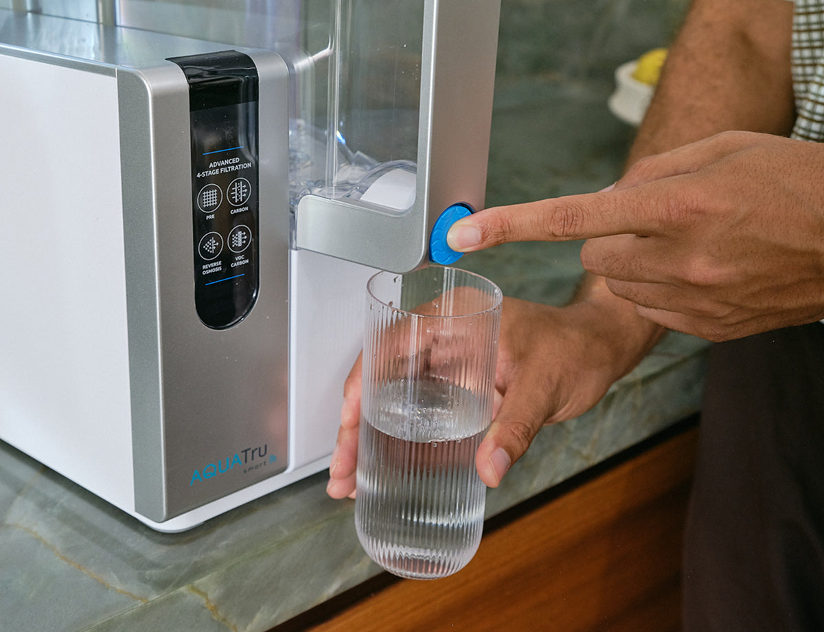 A hand fills a glass with water from the AQT Counter Classic water dispenser on a kitchen countertop.