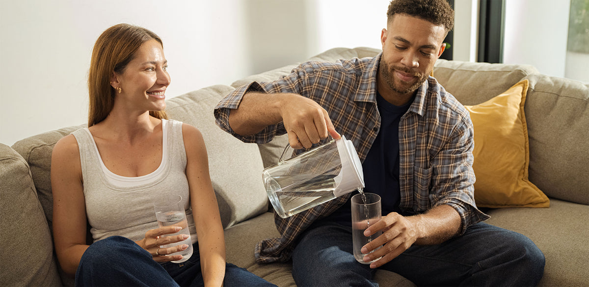 A man and woman sit on a couch in a living room setting. The man is pouring a glass of water from the AquaTru Glass Carafe.