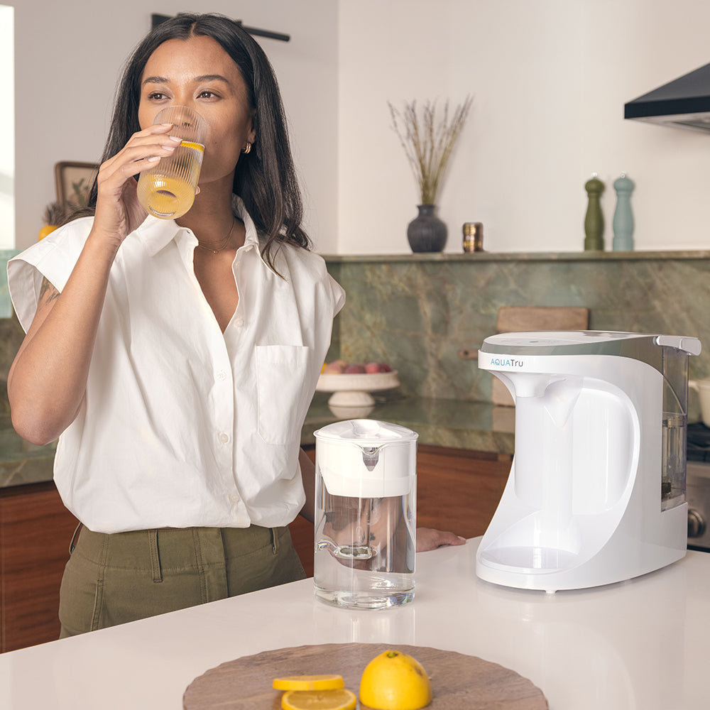 A woman stands at her kitchen island drinking a glass of water with a lemon slice that was purified with her AquaTru Carafe.