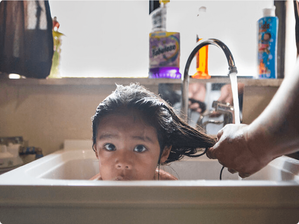 A child with wet hair looks curiously at the camera while someone washes their hair in a sink.