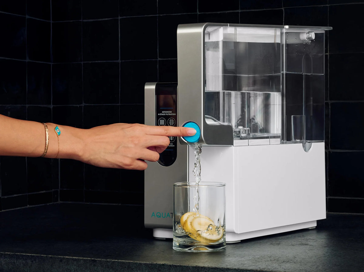 A hand presses a button on a water dispenser, filling a glass with water and lemon slices.