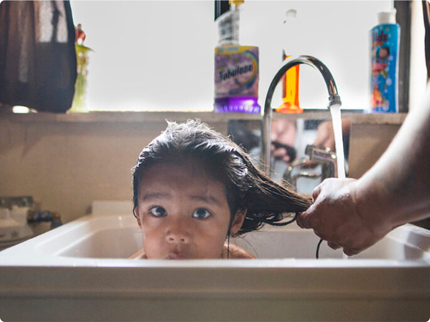 Child in a sink getting their hair washed, with colorful bottles in the background.