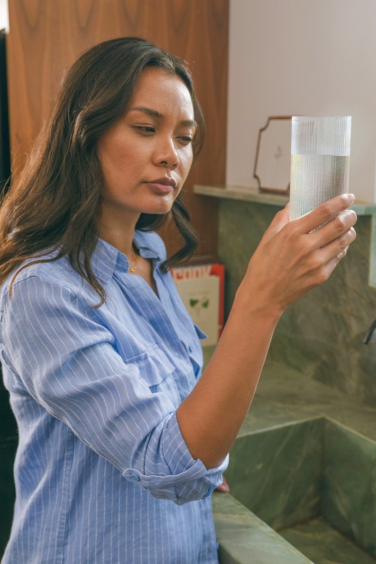A woman with long dark hair wearing a light blue and white striped button-down shirt holds up a clear glass tumbler with a white patterned insert, set against a bathroom sink background.