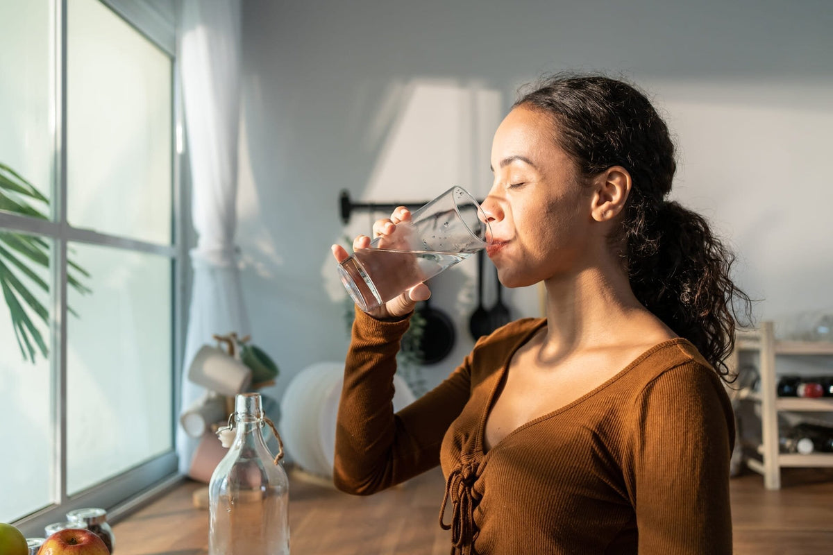 A woman drinks from a clear glass of water in a sunlit kitchen.