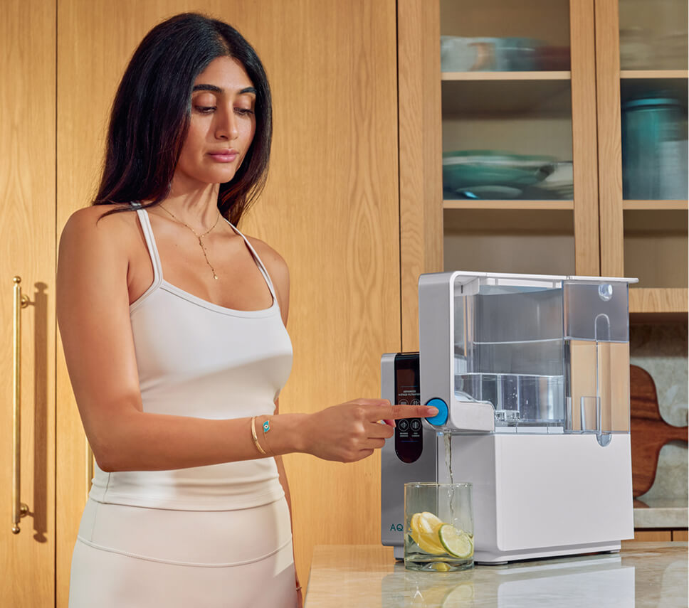 Woman in a white tank top using a water filtration system in a modern kitchen, with a glass of lemon-infused water nearby.