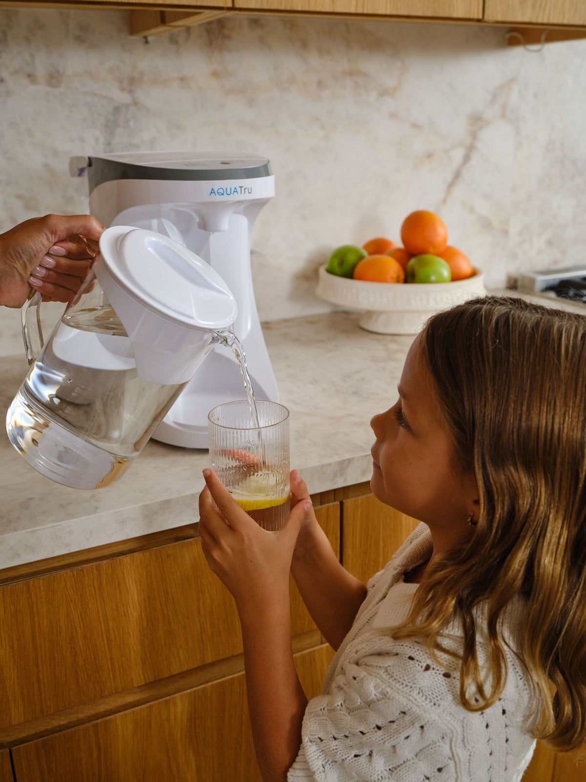 In a kitchen setting, a woman's hand is shown pouring water from an AquaTru Glass Carafe into a glass for a little girl. The AquaTru Carafe water purifier is on the countertop. 