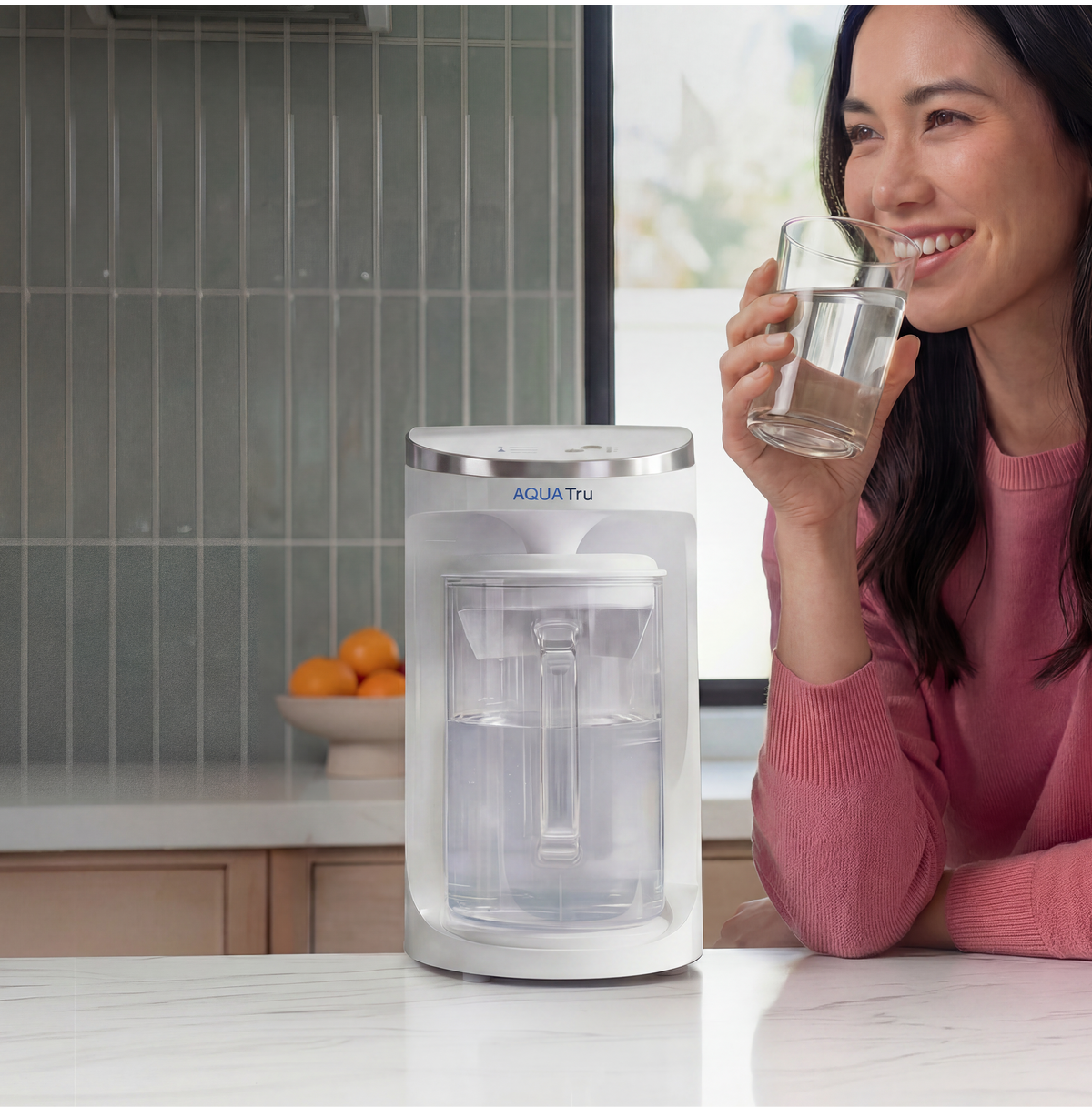 A woman smiles while holding a glass of water next to a sleek water filtration carafe on a kitchen counter.