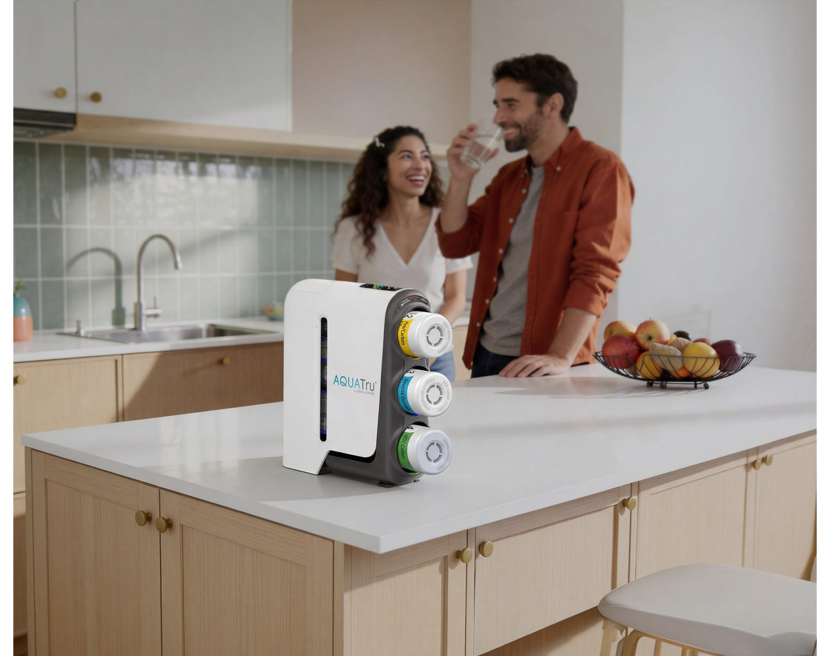 A couple enjoys drinks at a kitchen counter with a stylish beverage dispenser and a fruit basket in the background.