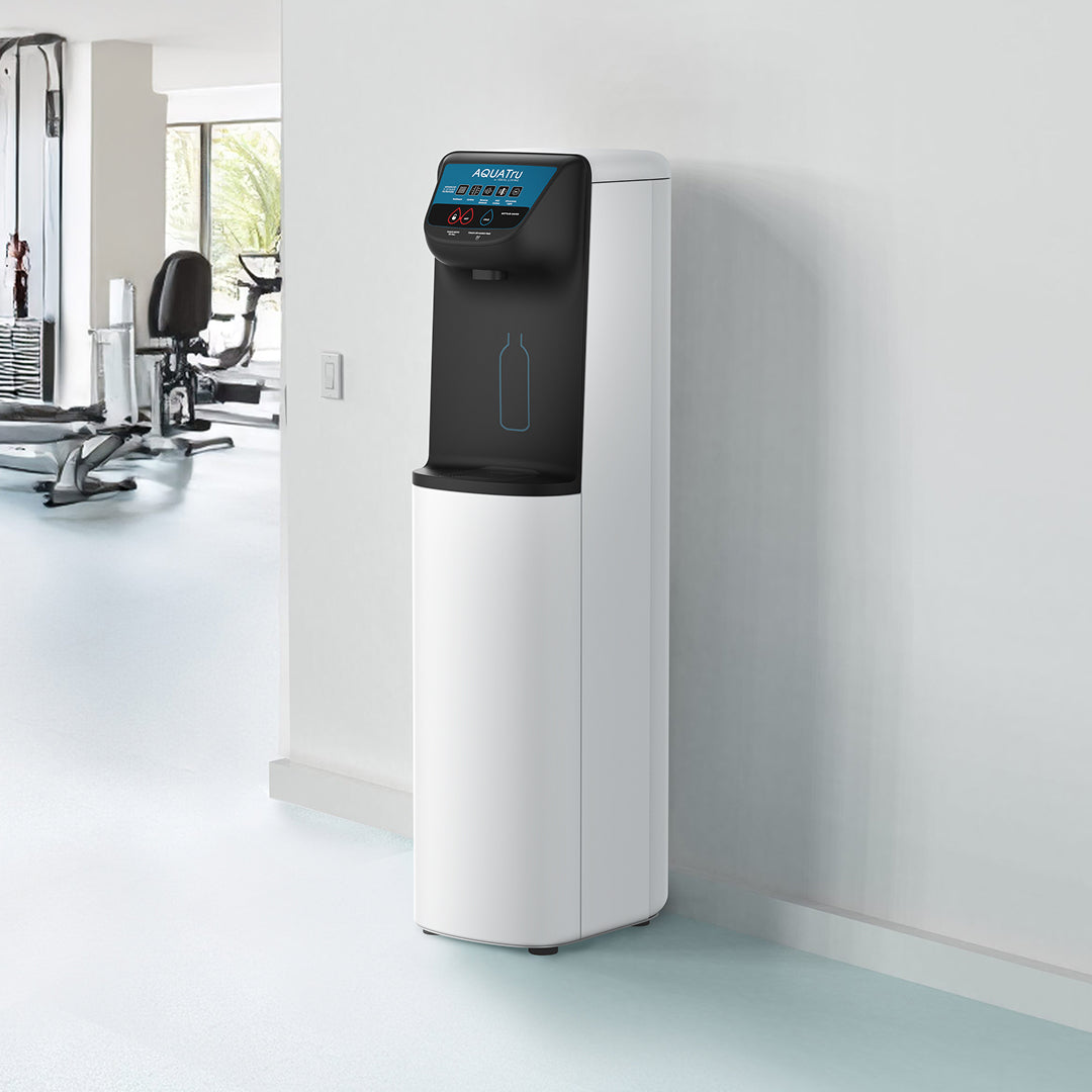 Water dispenser with a digital display, positioned in a bright gym environment with exercise equipment in the background.