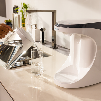 AquaTru Carafe pouring water into a glass next to a sleek white water purifier on a modern kitchen counter.
