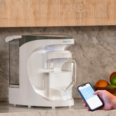 AquaTru carafe on a countertop with a hand holding a smartphone, surrounded by fruits in the background.