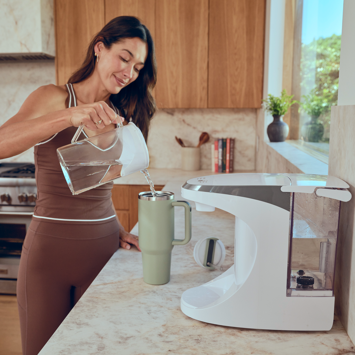 A woman pours water from a pitcher into a large mug next to a countertop water dispenser in a modern kitchen.