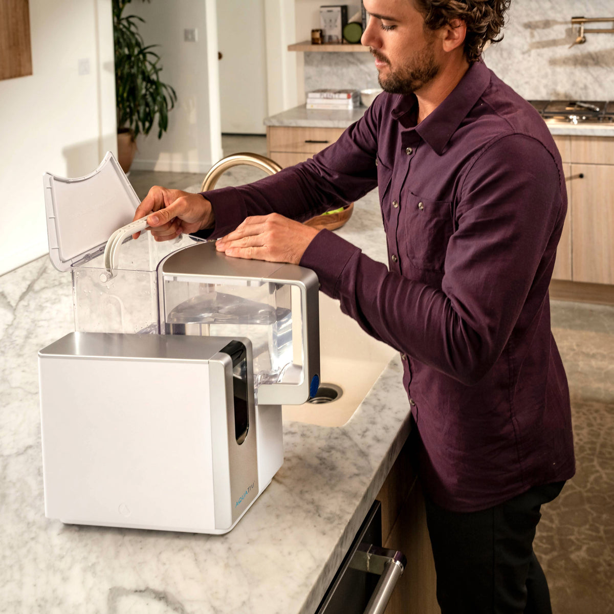 A man in a purple shirt fills a water filter pitcher in a modern kitchen with marble countertops.