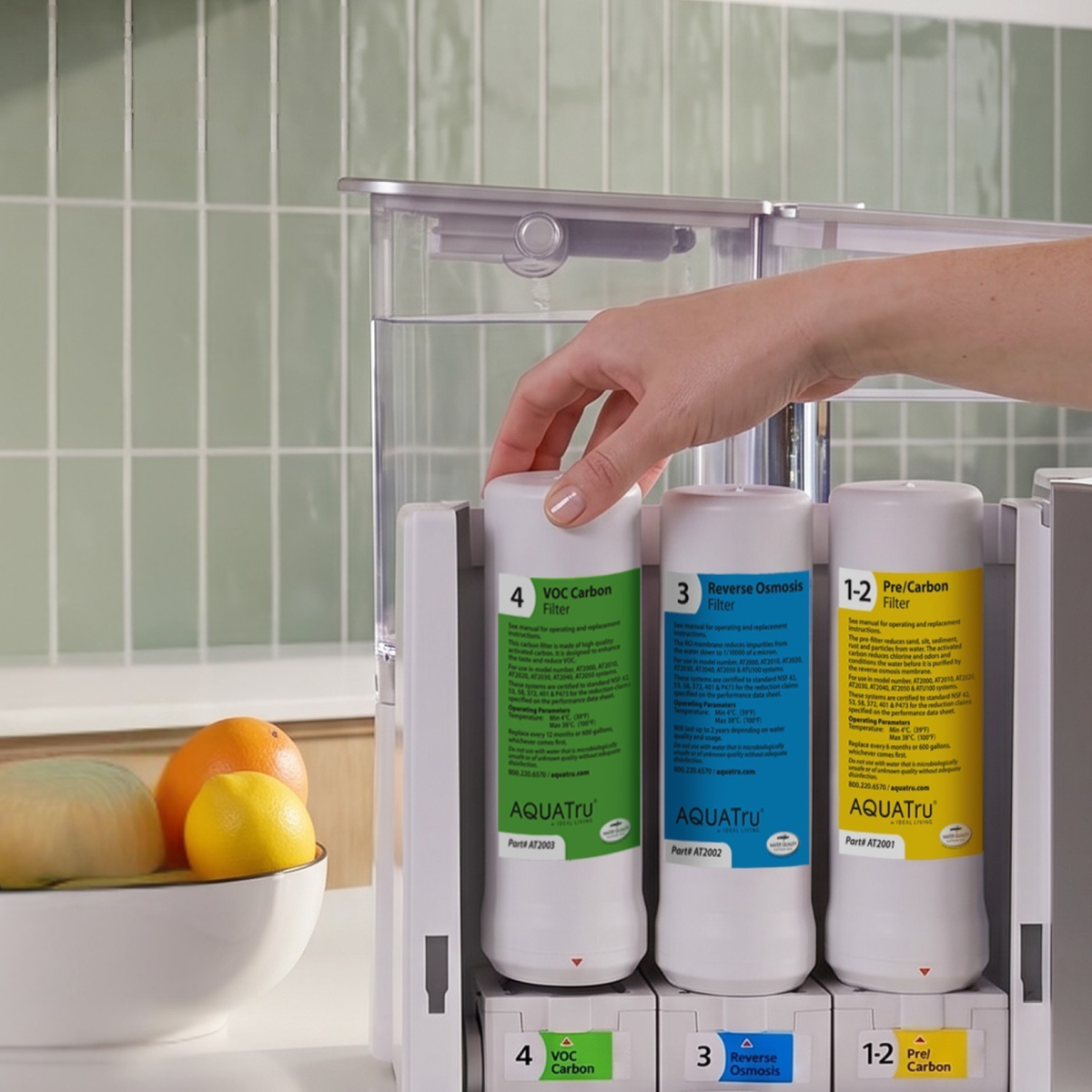 Hand placing a water filter into a countertop filtration system, with fresh fruits in a bowl nearby.