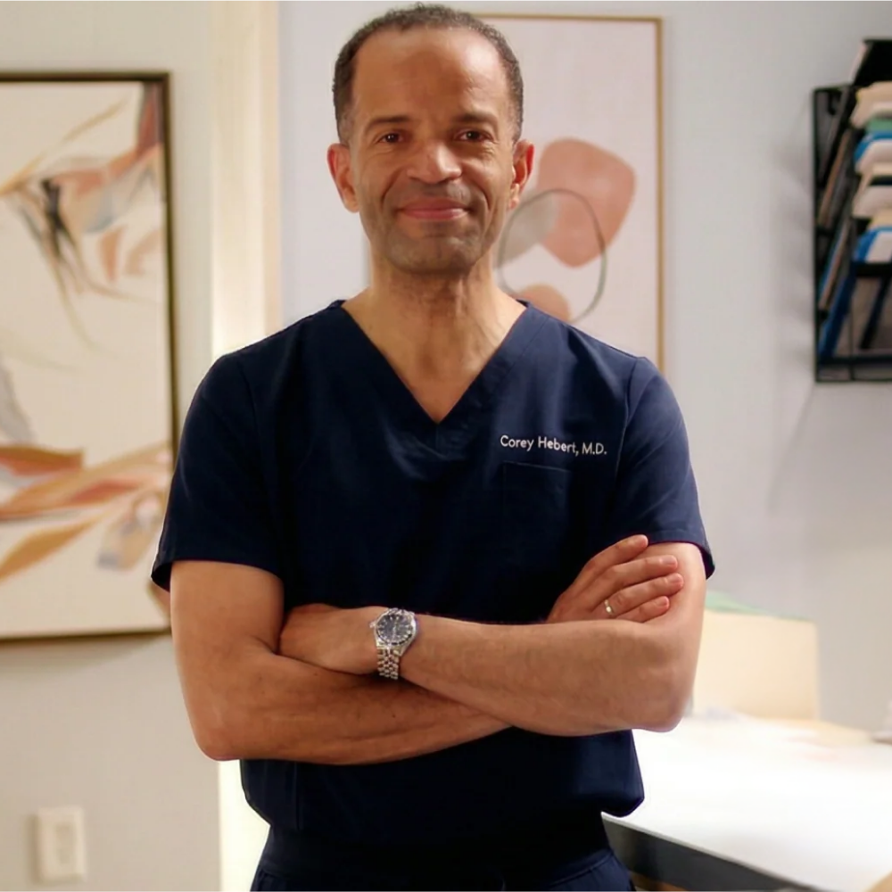 Dr. Corey Hebert, M.D., stands confidently with arms crossed, wearing a navy scrub top in a clinical setting.