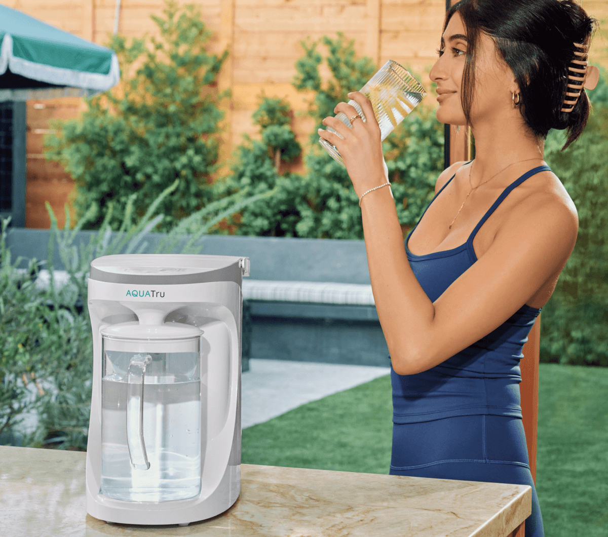 A woman in a blue tank top drinks water from a glass beside a white AQUA Tru water purifier on a marble countertop.
