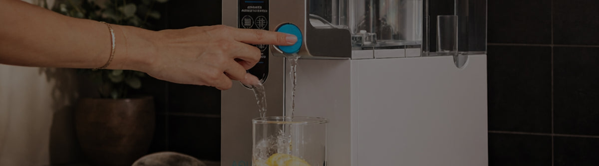 A hand presses a blue button on a water dispenser, filling a glass with water and lemon slices.