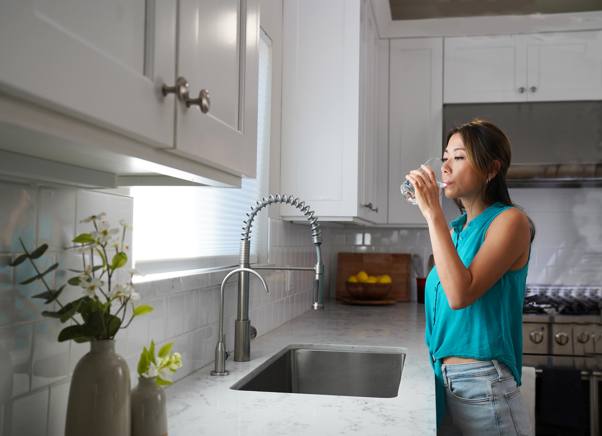 A woman drinks water from a glass at a modern kitchen sink, with plants and a bowl of lemons nearby.