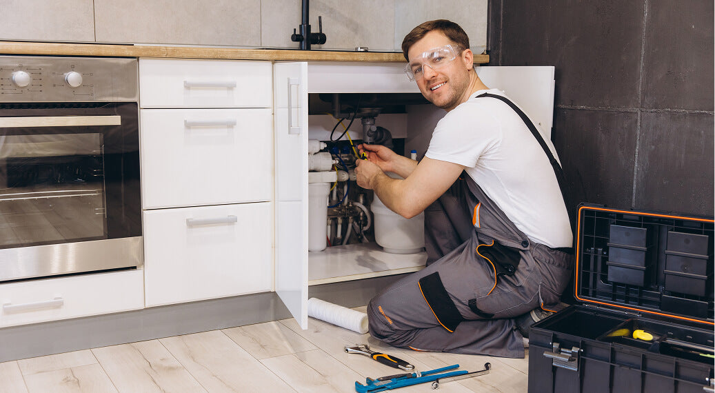 Plumber working under a kitchen sink, wearing safety goggles and holding tools, with a toolbox nearby.