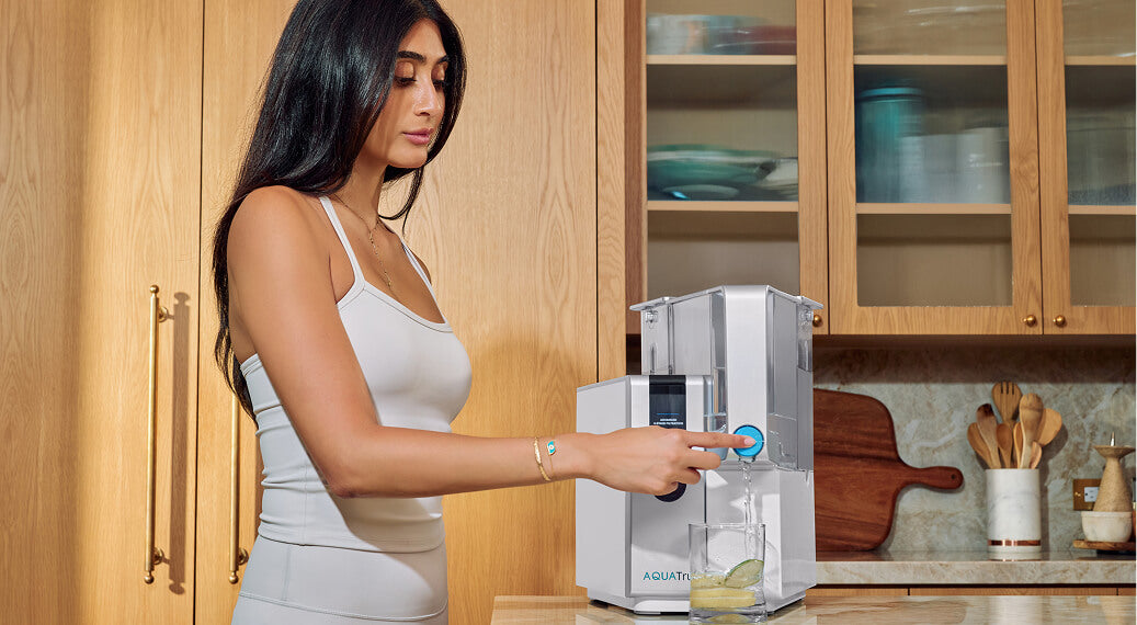 A woman in a white tank top uses a water filtration system in a modern kitchen, pouring water into a glass.