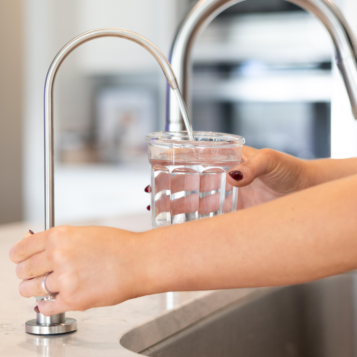 A person fills a clear glass with water from a modern kitchen faucet.