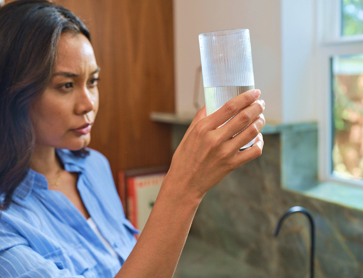 Woman examining a clear glass of water in a kitchen, with a focused expression and natural light illuminating the scene.