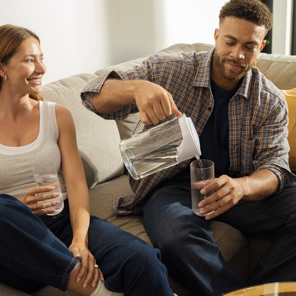 A woman and a man sit on a couch. The man is pouring water into a glass from the AquaTru Glass Carafe. 