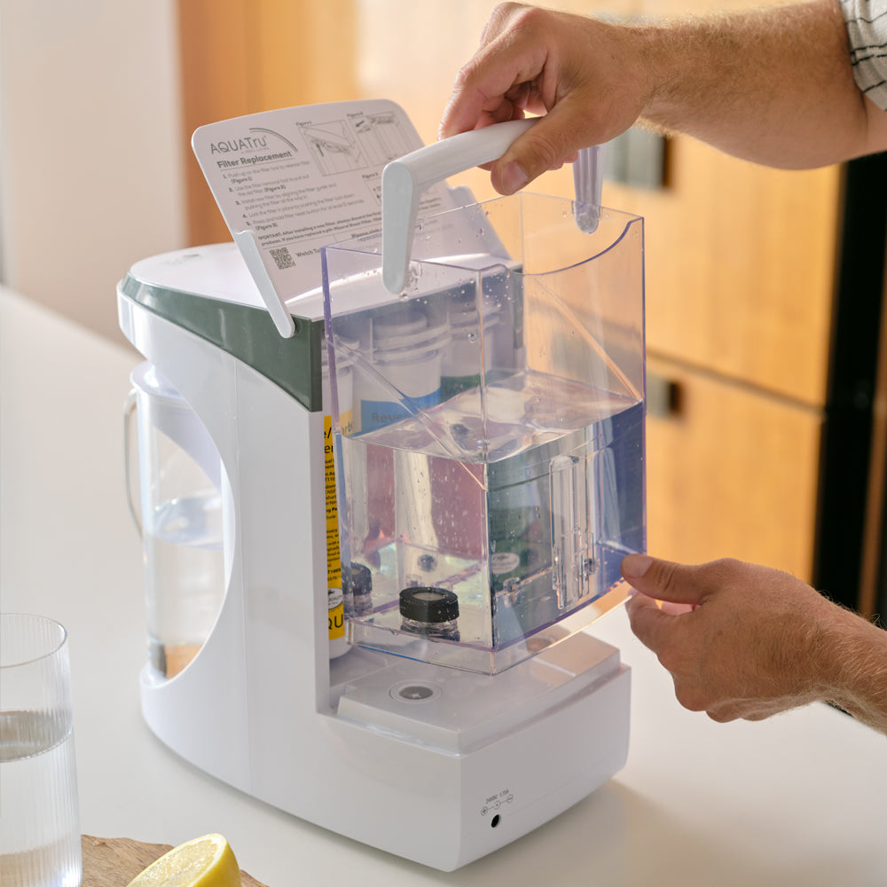 A man's hands are shown installing the AquaTru tap water tank to the back of the AquaTru Carafe. 