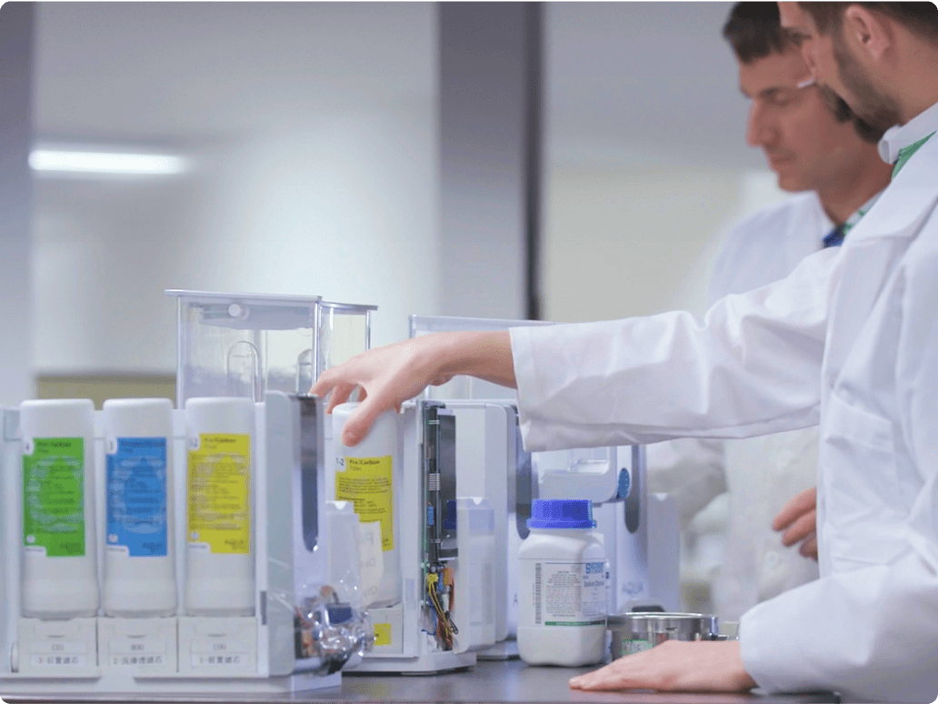 Two lab technicians in white coats operate equipment and examine containers in a laboratory setting.