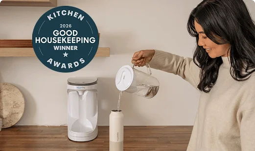 The image shows a woman pouring water purified through the AquaTru system from a glass carafe pitcher into an AquaTru Stainless bottle, featuring a 2026 Good Housekeeping Kitchen Awards Winner badge.