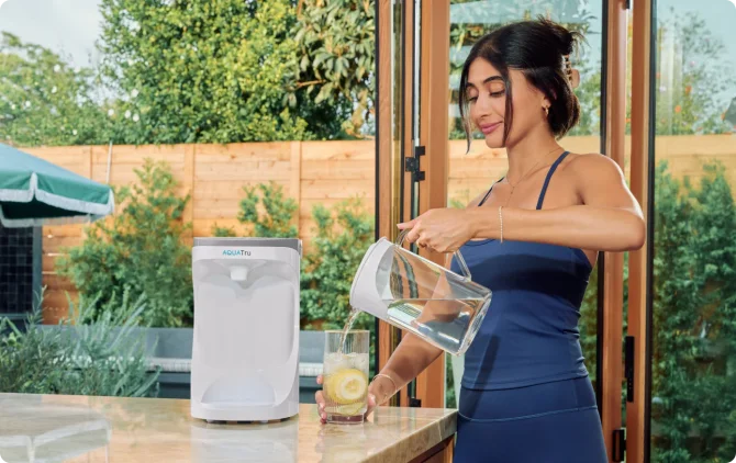 A woman in blue activewear pours water purified through the AquaTru system from a glass carafe pitcher into a glass.