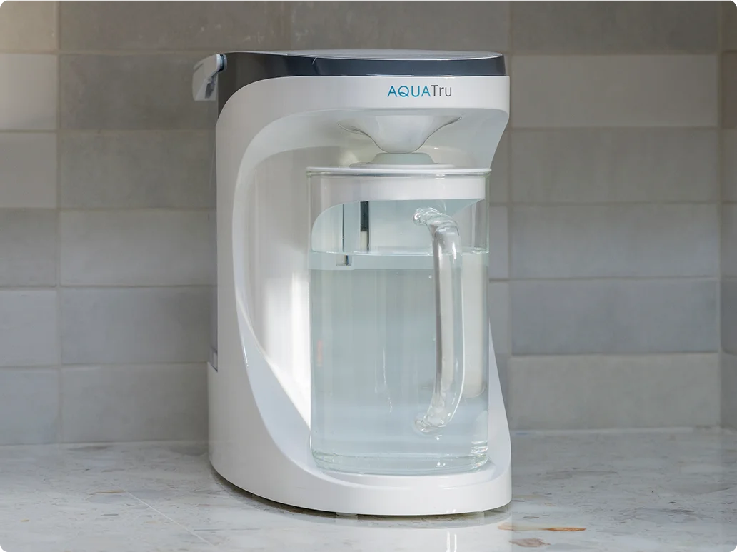 AQUAtru water filtration system with a clear carafe on a marble countertop against a tiled background.