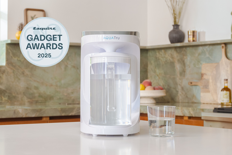 A white Aquatu water purifier sits on a kitchen counter next to a glass of water, with a 'Esquire Gadget Awards 2025' badge on the left. The background shows a blurred kitchen setting with green marble backsplash.