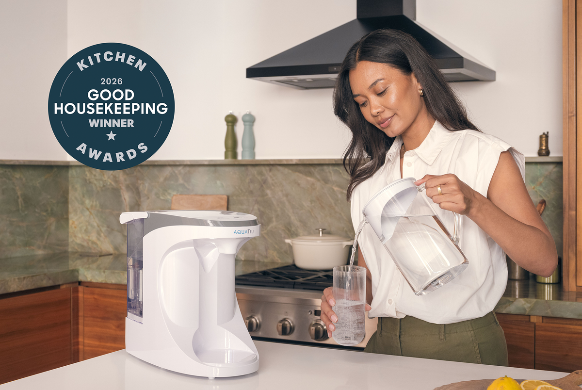 A woman in a white sleeveless blouse and olive pants pours water from a clear pitcher into a glass on a kitchen counter, with a white countertop water purifier beside her. A dark blue circular badge in the upper left reads 'Kitchen 2026 Good Housekeeping Winner Awards.