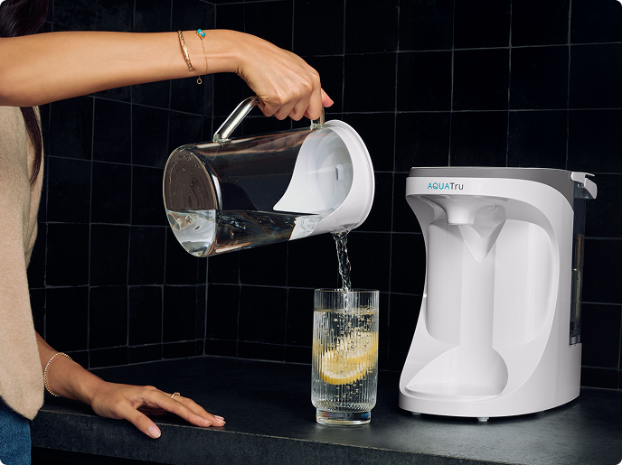 A person pours water from a pitcher into a glass with lemon, next to a water purification system on a countertop.