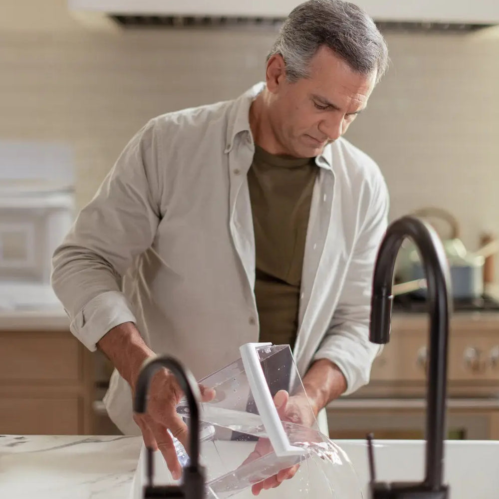 Man spills water from a clear container while standing at a kitchen counter, with modern faucets in the foreground.