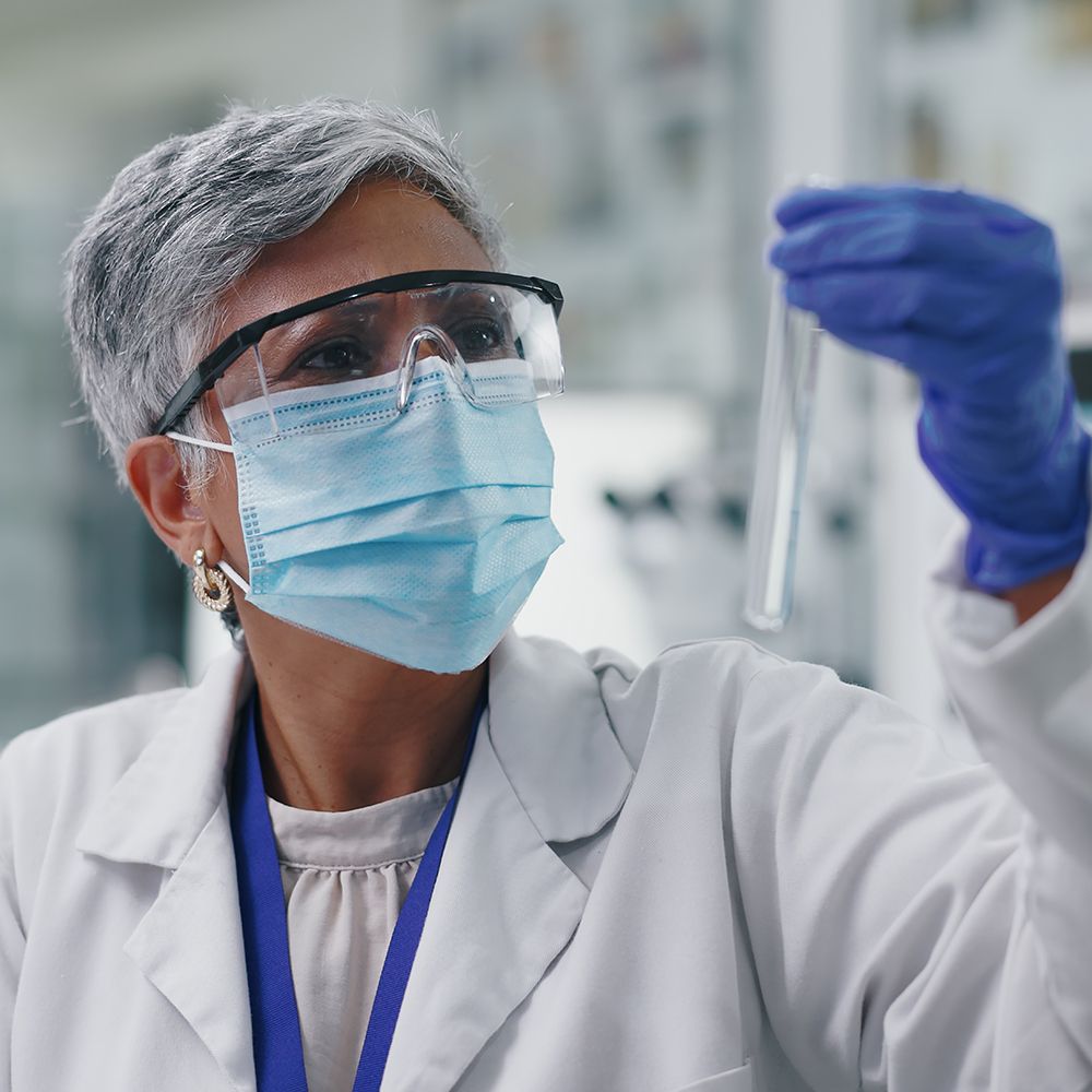 A close-up of a scientist or doctor wearing a white lab coat, light blue face mask, clear safety glasses, and blue nitrile gloves, holding a test tube up in a laboratory setting.