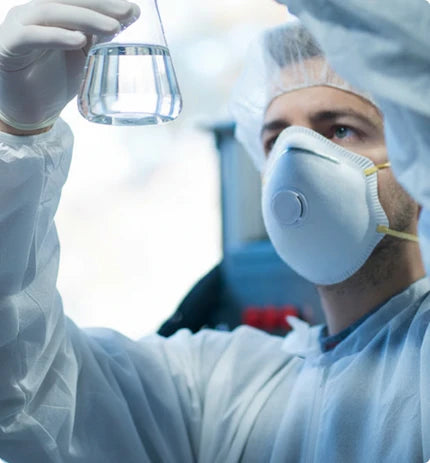 A laboratory scientist in full protective gear examines a glass flask filled with water for safety testing.