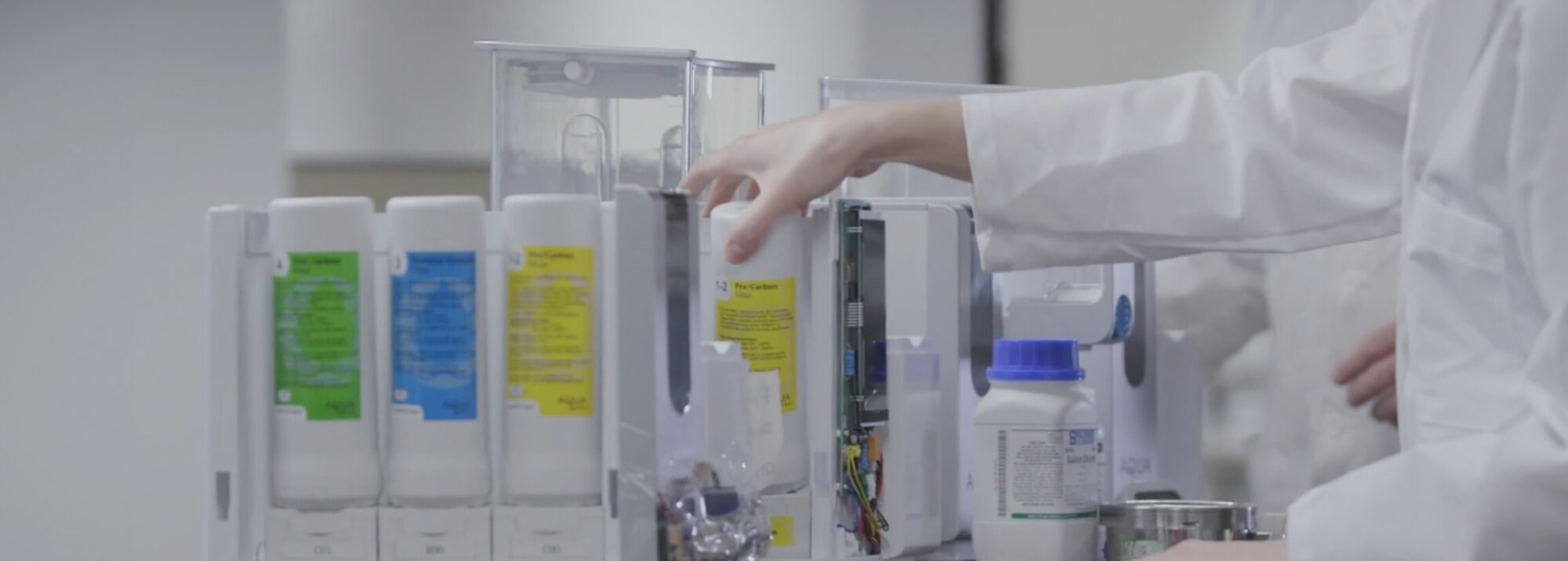 A laboratory setting shows a technician in a lab coat inserting a yellow filter cartridge into an AquaTru water purification unit.