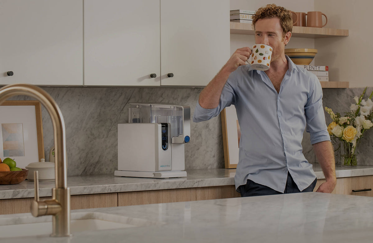 Man in a light blue shirt sipping from a patterned mug next to a water dispenser in a modern kitchen.
